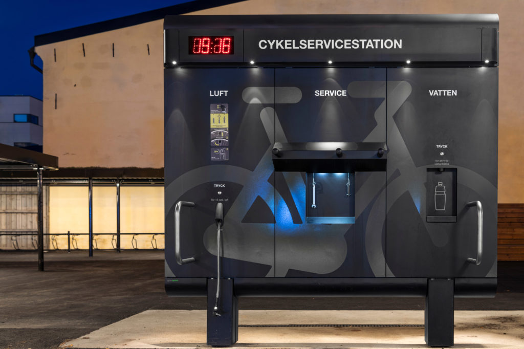 Cykelservicestation GRAND från Cyklos i skymningsljus, med LED-belysning, cykelpump, multiverktyg, påfyllning av vattenflaska och på baksidan en tvätt.