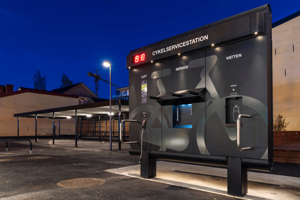 Cykelservicestation GRAND från Cyklos i skymning med LED-belysning, cykelpump, multiverktyg, påfyllning av vattenflaska och på baksidan en tvätt.
